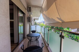 Un balcón con mesa y sillas, unidades de aire acondicionado y vistas a zonas verdes y edificios.