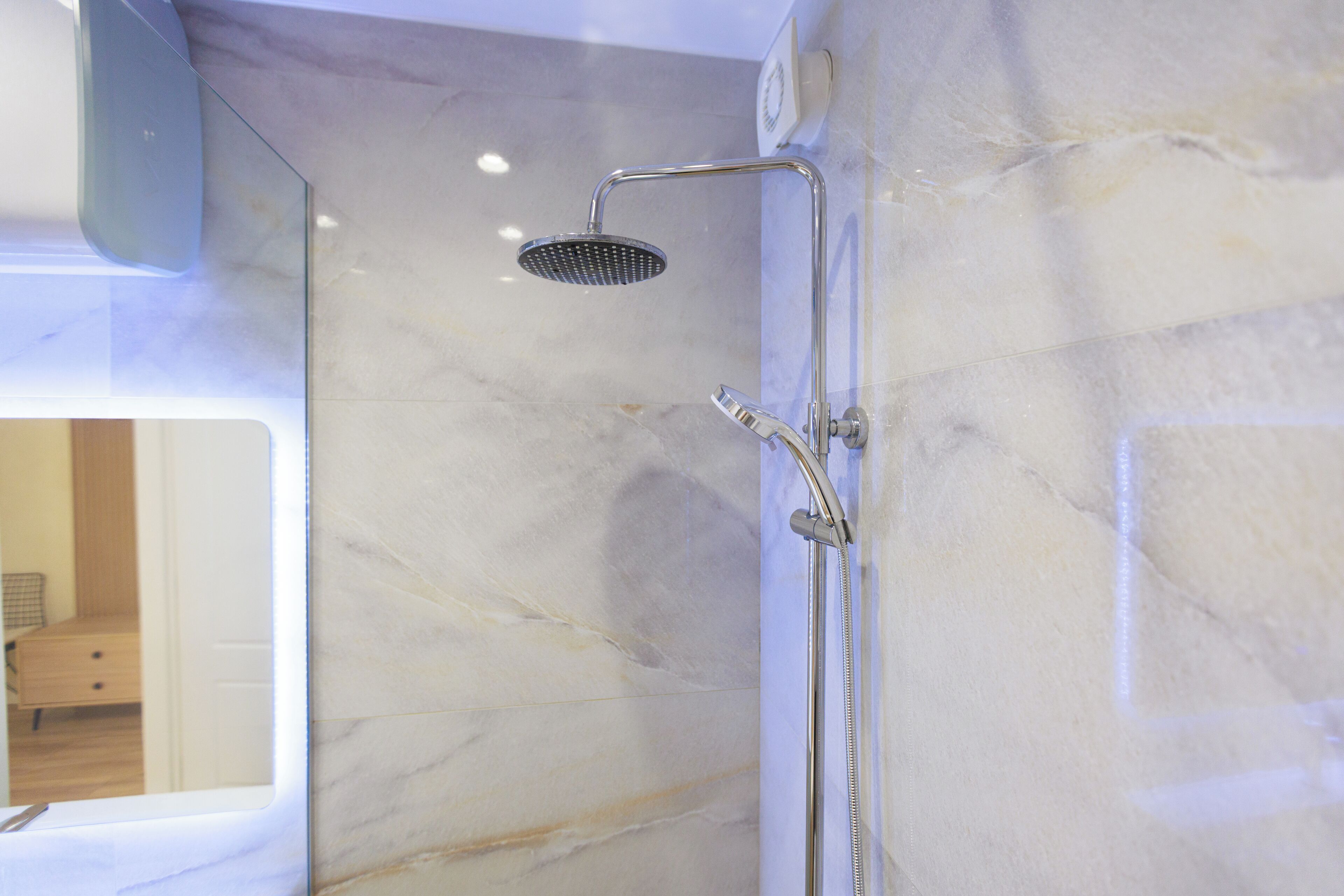 A modern bathroom with a shower, marble walls, and a visible bedroom with a bed and dresser.