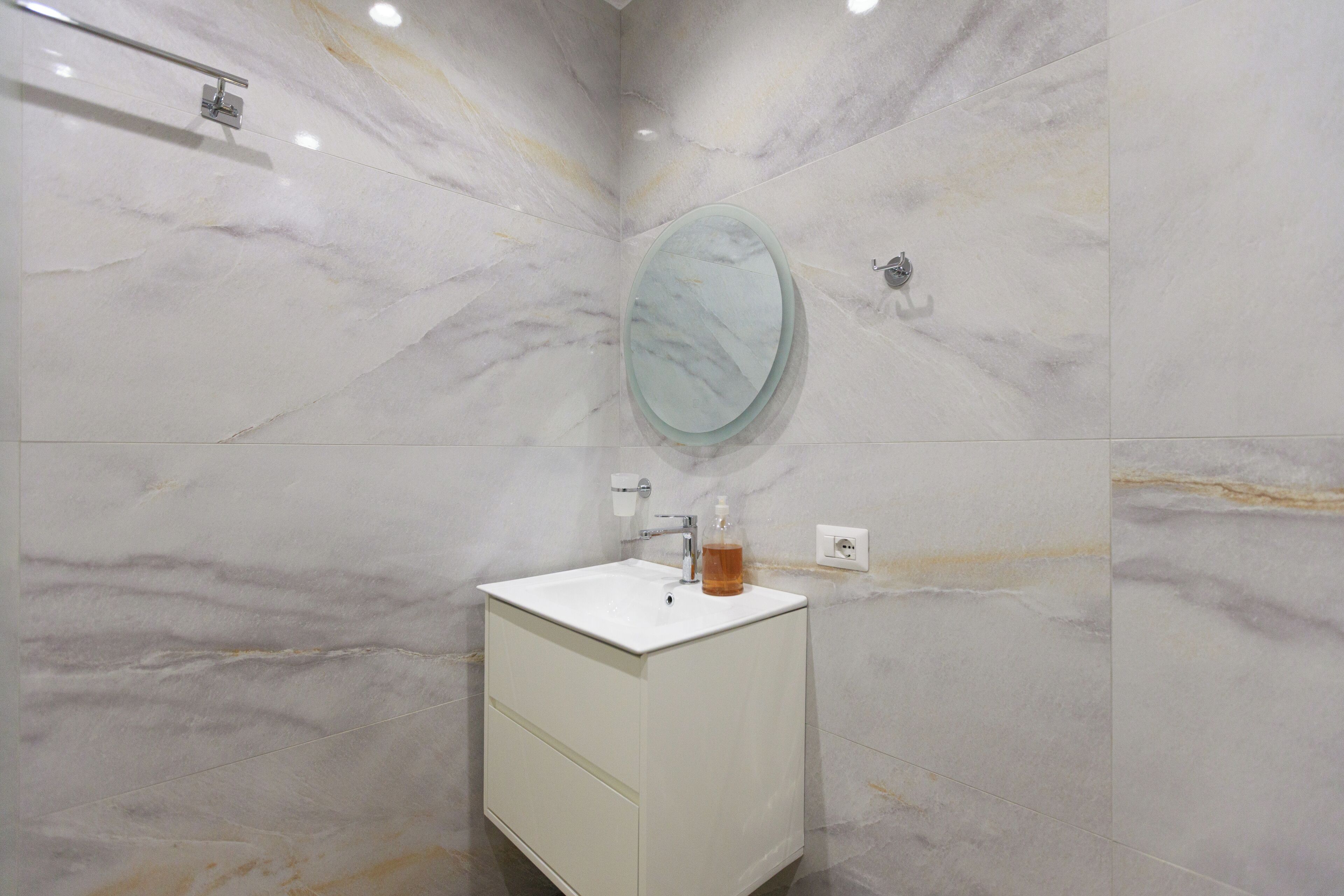 Une salle de bain avec un mur en marbre, un miroir rond, un lavabo blanc et un distributeur de savon.