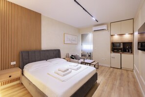Moderni hotellihuone, jossa on suuri sänky, pieni työpöytä, tuoli, sohva, pieni keittiö ja ilmastointilaite.