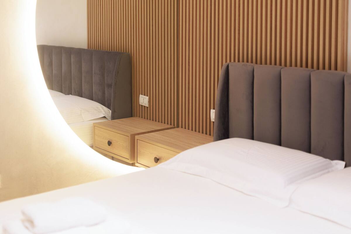 Een moderne hotelkamer met een grote ronde spiegel, een bed met witte lakens, een houten wandpaneel en een nachtkastje met een lamp.