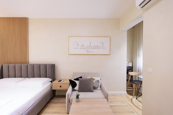 Un dormitorio moderno con cama, mesita de noche y un pequeño sofá. En la pared, un cuadro enmarcado con una vista urbana.