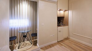 Een compacte kamer met een kitchenette, een kleine eethoek met een tafel en twee stoelen, en een raam met gordijnen.