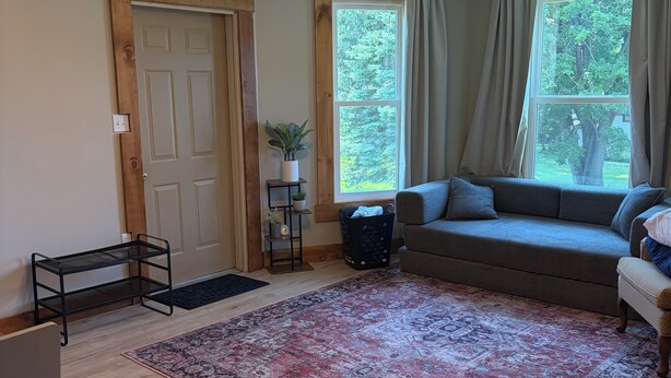 A living room with a wooden floor, a large window with curtains, a sofa, a rug, and a ceiling light fixture.