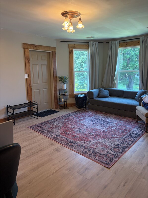 A living room with a wooden floor, a large window with curtains, a sofa, a rug, and a ceiling light fixture.