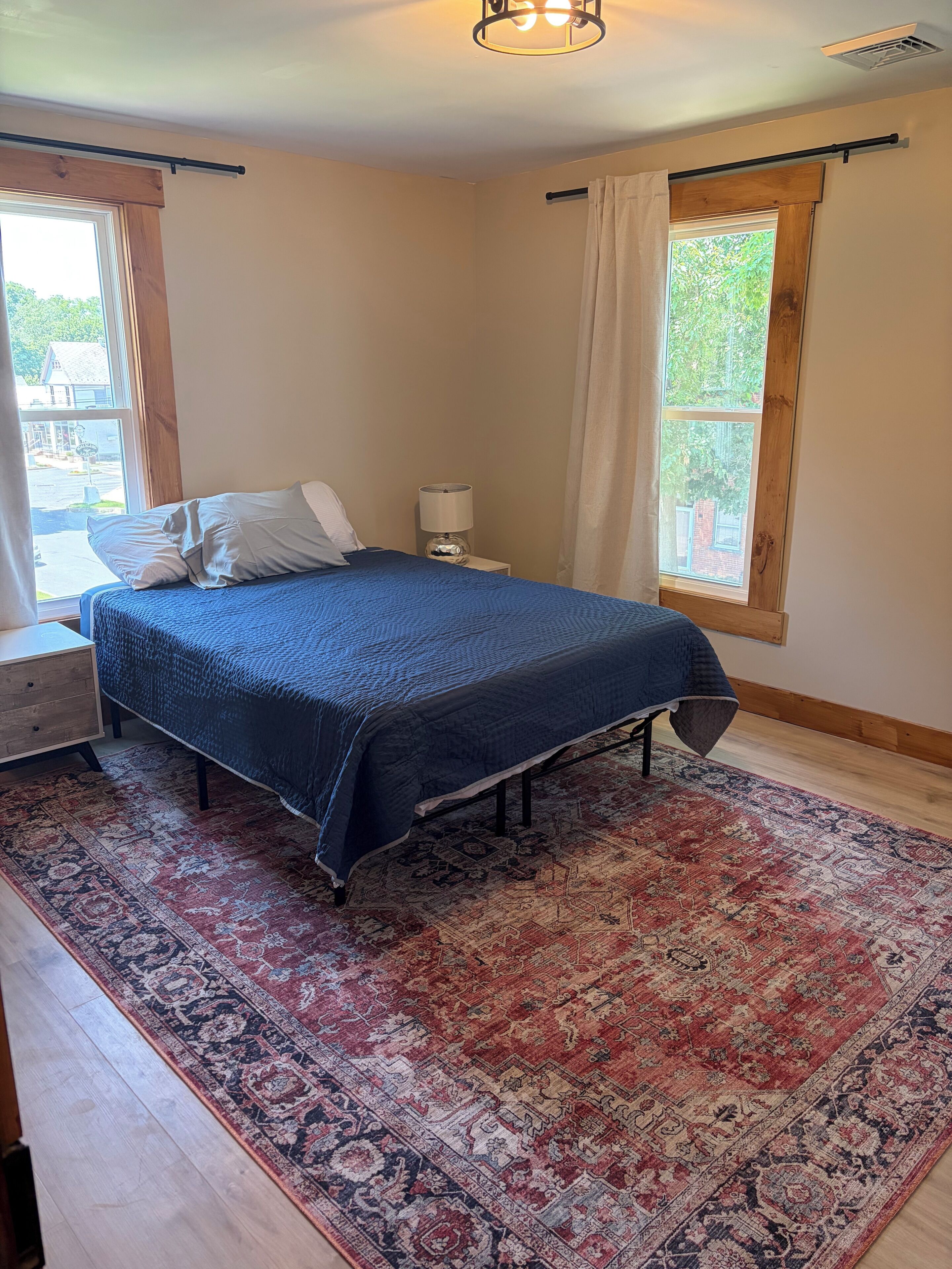 A bedroom with a bed, a nightstand, a lamp, a window with curtains, and a patterned rug.