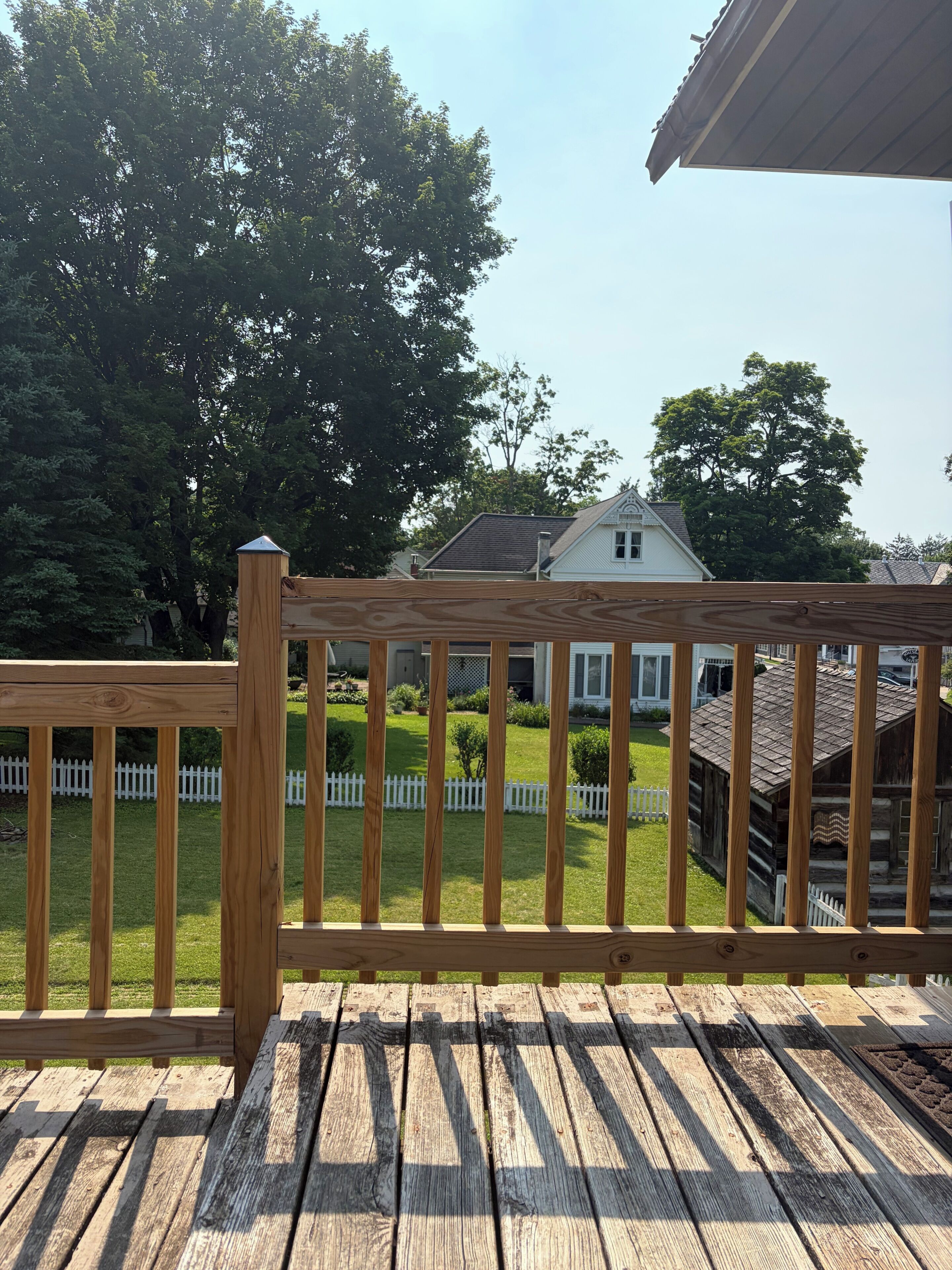 A wooden deck with a white picket fence, a grassy yard, and a house with a sloped roof in the background.