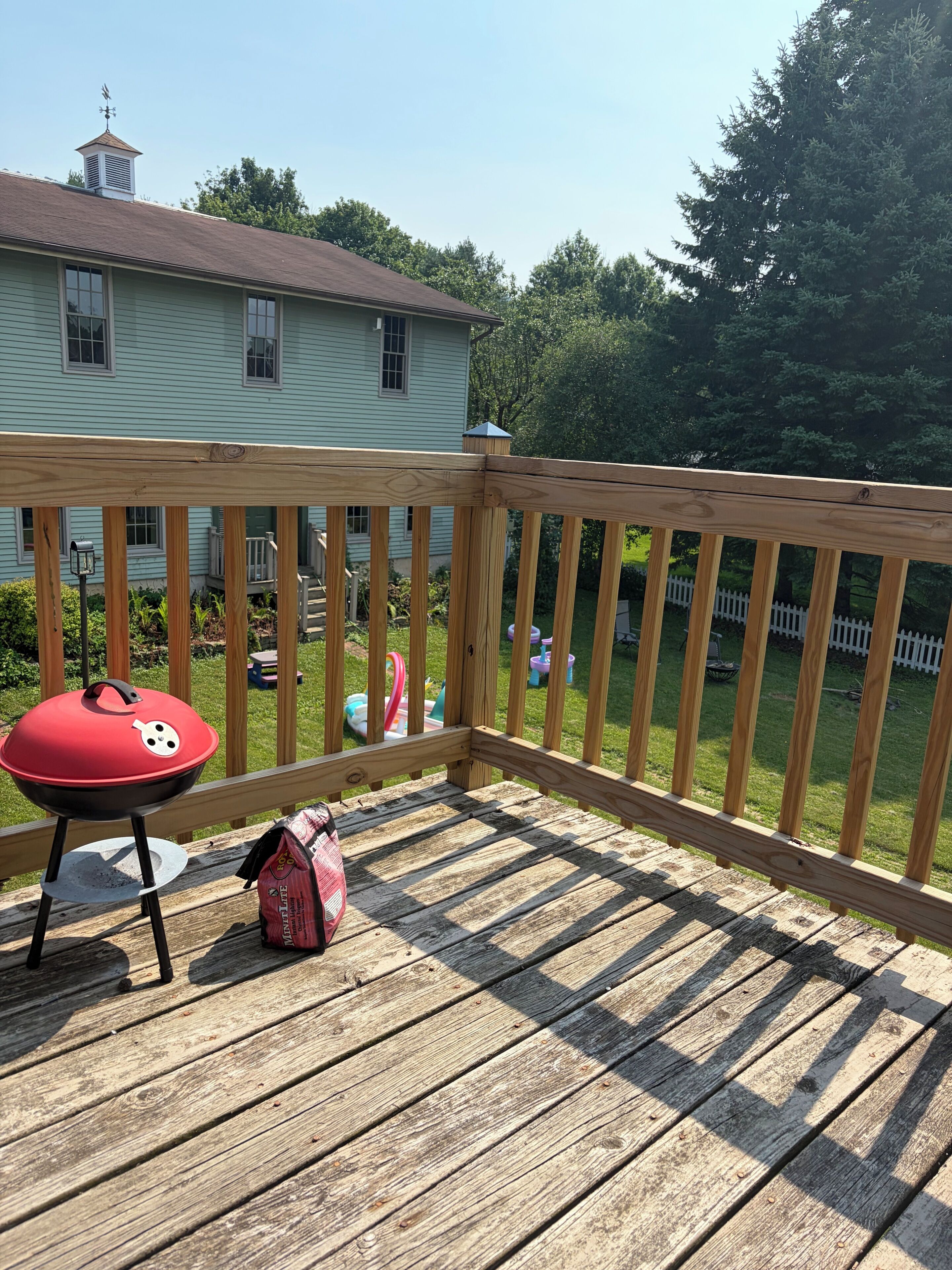 Una terraza de madera con una parrilla roja y bolsas de carbón, con vista a un patio trasero con cerca blanca y follaje.