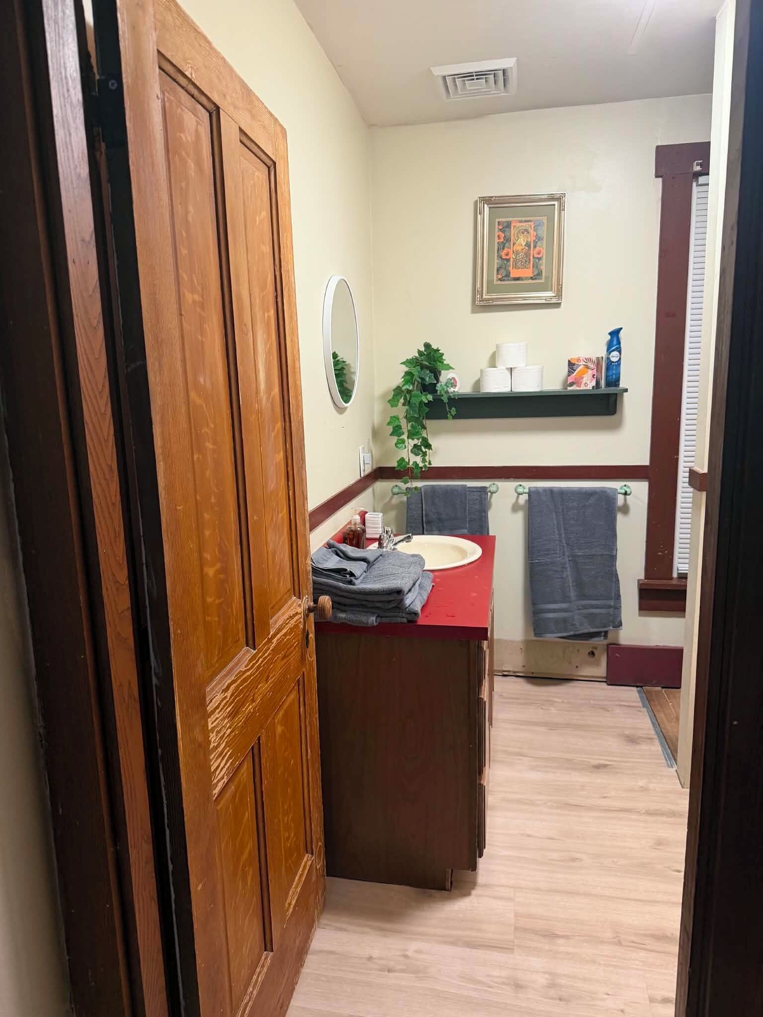 Ein Badezimmer mit Holztür, Waschbecken in Rot, Spiegel, Handtuchhalter und Ablagefläche für Toilettenartikel.