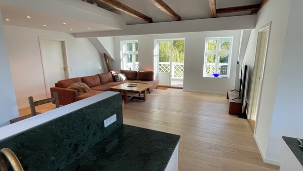 A spacious living room with a wooden ceiling, a brown leather sofa, a wooden coffee table, and a large window with a view of greenery.