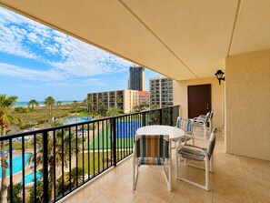 A balcony with a view of a swimming pool and palm trees, a table with chairs, and a lamp. - Three Bedroom Two Bath Beachfront Condo (South Padre Island)