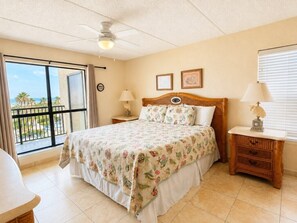 A bedroom with a bed, two bedside tables, a ceiling fan, a window with a view, and a door leading to an outdoor balcony. - Three Bedroom Two Bath Beachfront Condo (South Padre Island)