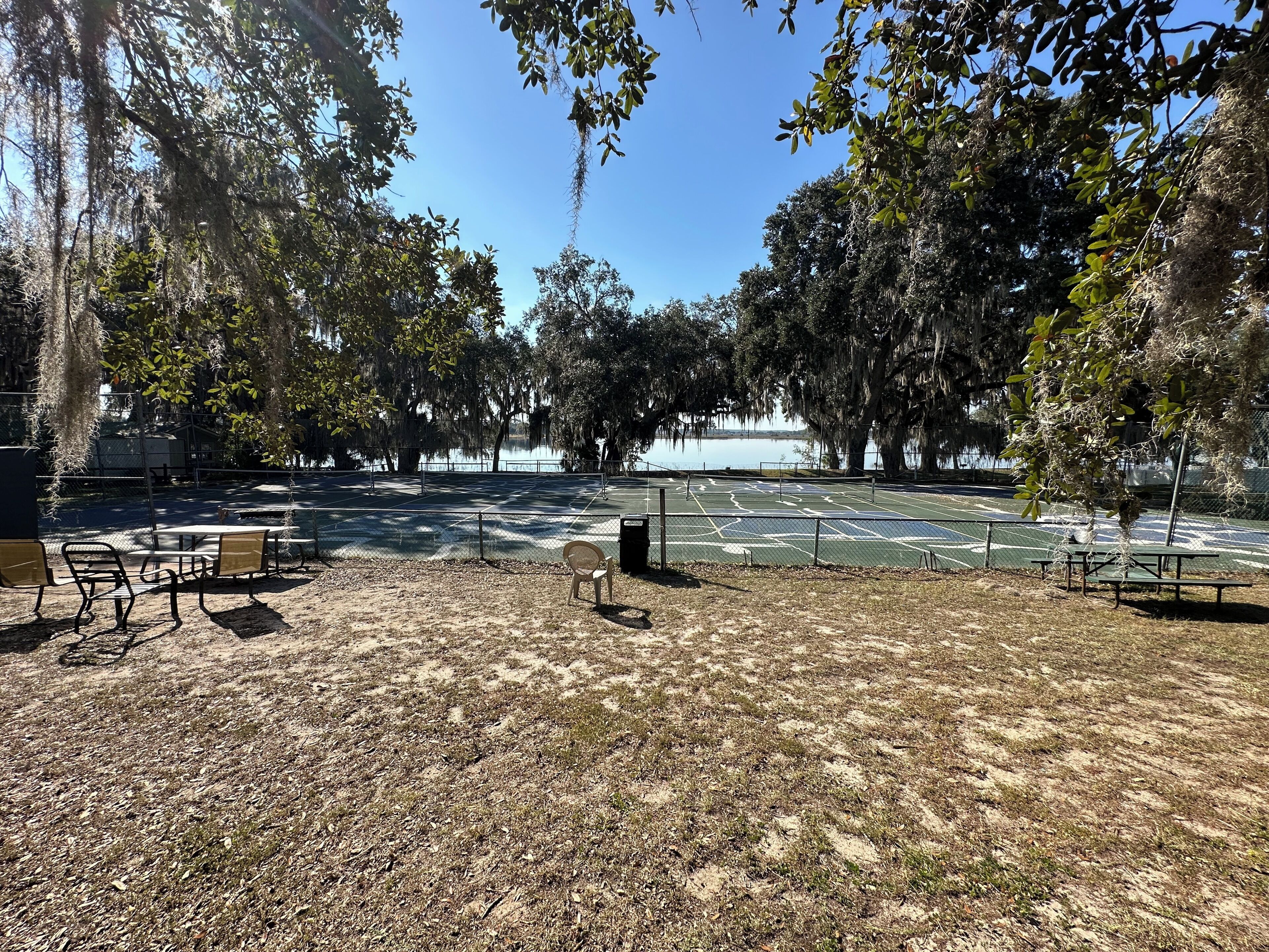 An outdoor tennis court with benches and picnic tables, surrounded by trees and a body of water.