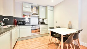 A modern kitchen with white cabinets, a built-in oven, and a dining area with a white table and black chairs. - Luxury One-Bedroom by Tower of London • Prime Location (London)