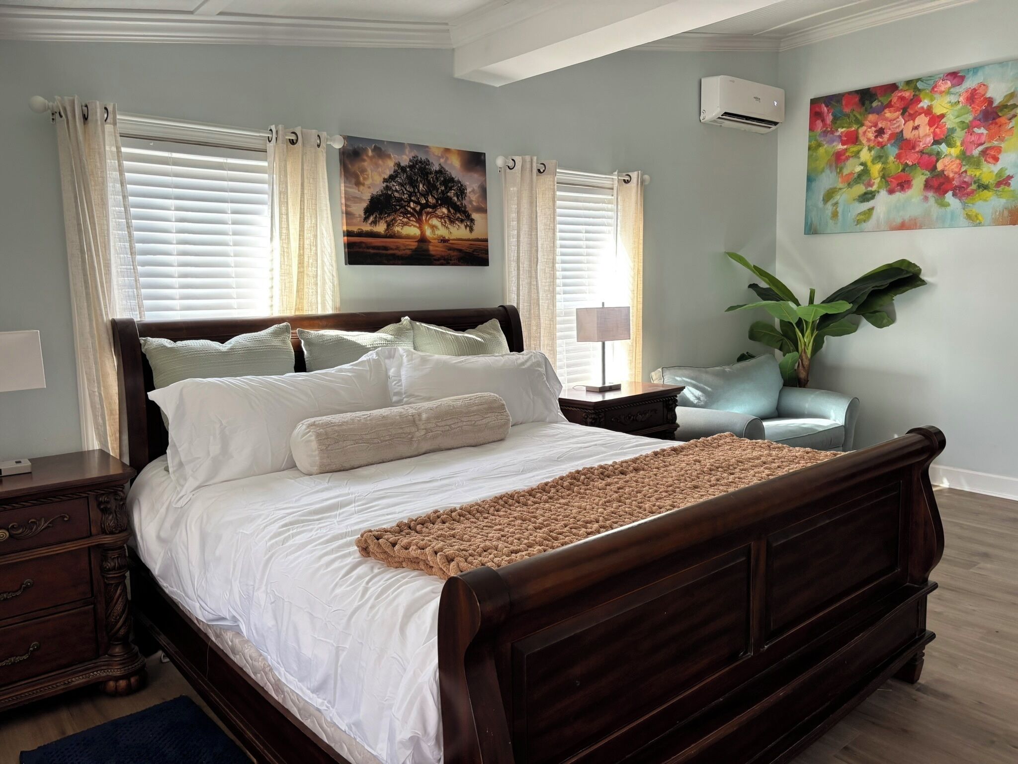 A bedroom with a wooden bed, white bedding, a decorative throw, and a painting of a tree.