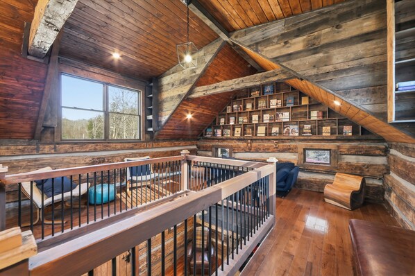 Het interieur van een blokhut met een houten trap, een boekenkast en uitzicht op de natuur.