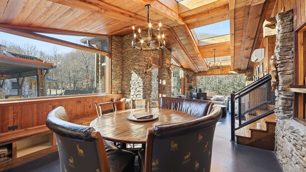 A wooden dining area with a stone wall, a chandelier, and a view of the outdoors. - Lodge at Wagon Gap Trl. 3 Blue Ridge River Cabins (Canton)
