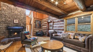 A cozy log cabin interior with a stone fireplace, leather furniture, and a large window. - Lodge at Wagon Gap Trl. 3 Blue Ridge River Cabins (Canton)