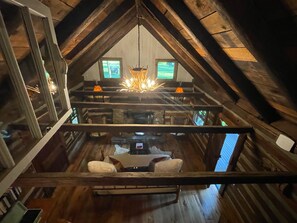 An attic room with a sloped ceiling, wooden beams, a chandelier, two computer monitors, and a cozy seating area with cushions.