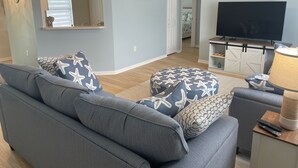 A living room with a sofa, ottoman, and a television. There is a door leading to another room. - Private Heated Screened Pool,  10 min to Beach, Coastal, Comfy, 3Bd/2B, Quiet
 (Sebastian)