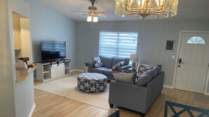 A living room with a grey sofa, ottoman, and a TV on a stand. There is a ceiling fan, a chandelier, and a door in the background. - Private Heated Screened Pool,  10 min to Beach, Coastal, Comfy, 3Bd/2B, Quiet
 (Sebastian)