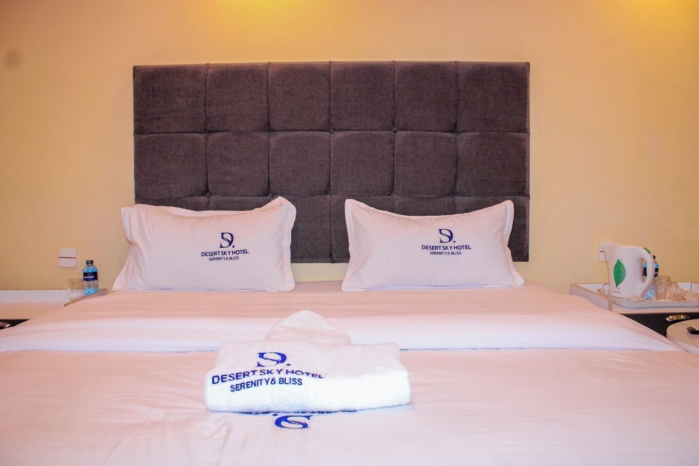 Een hotelkamer met een groot bed, twee kussens met het logo van het hotel, een opgevouwen handdoek en een nachtkastje met een waterkoker.