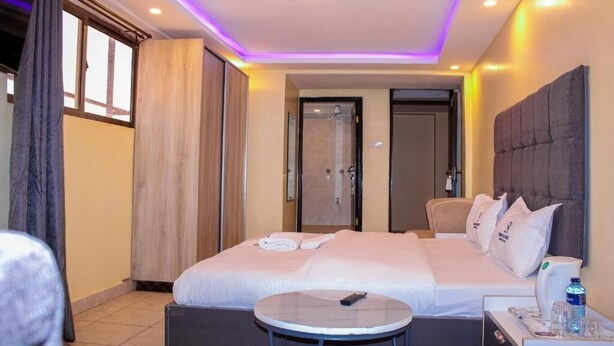 A hotel room with a large bed, a bedside table with a remote control, a small round table, and a bathroom door. - Desert Sky Hotel (Isiolo)