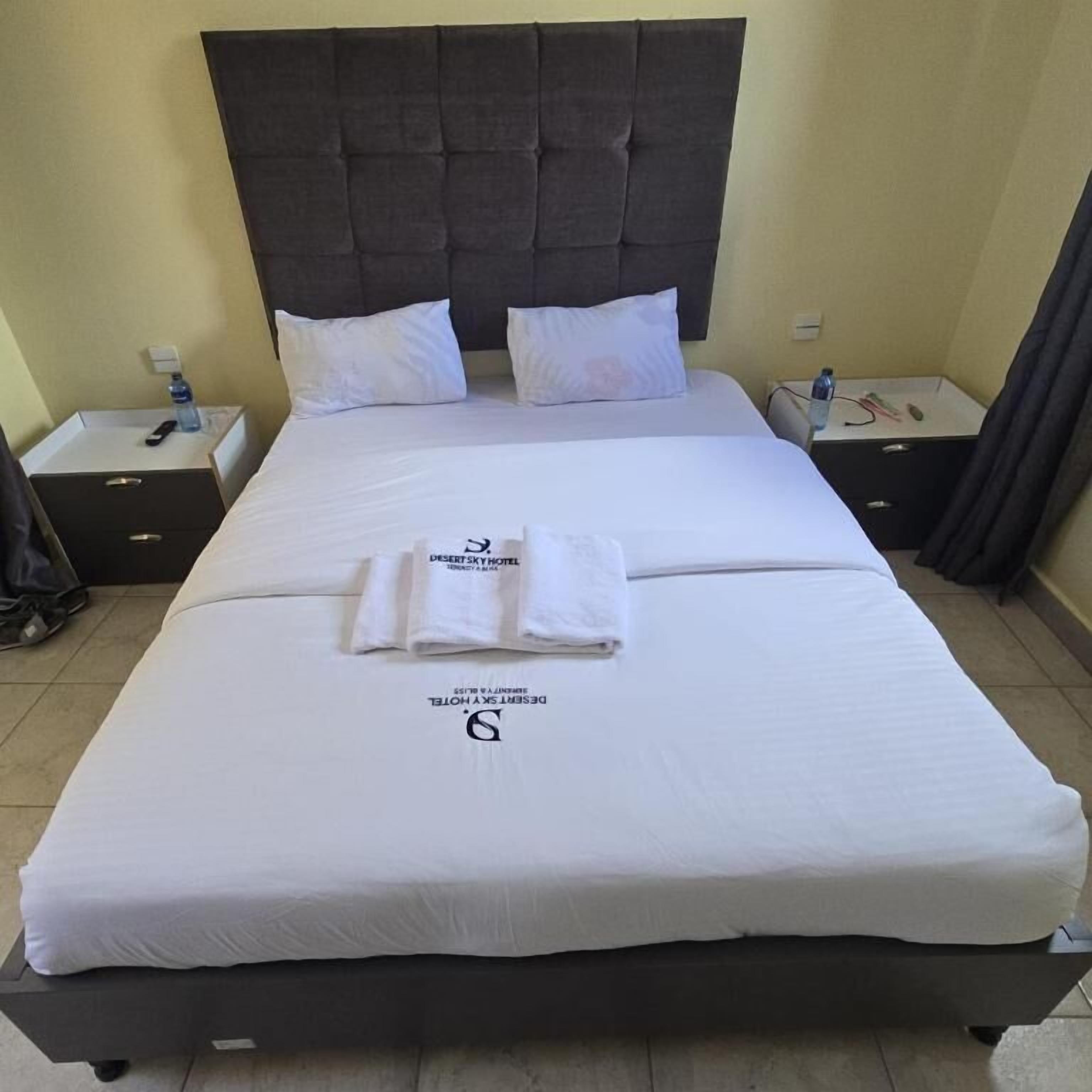 A neatly made bed with white linens, a dark headboard, and two pillows. There are two bedside tables, each with a remote control and a bottle of water. The room has a tiled floor and a plain wall.