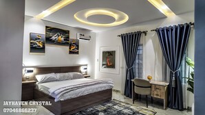 A modern bedroom with a large bed, a desk, and a chair. There are ceiling lights, artwork on the walls, and a window with curtains. - Jayhaven Crystal Serviced  Apartment-One Uyo (Uyo)