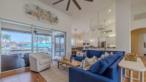 A spacious living room with a blue sofa, white armchair, and a coffee table. A large window offers a view of palm trees and a pool. - Moondance Villa (Cape Coral)