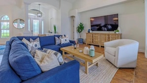 A spacious living room with a blue sofa, a white armchair, a wooden coffee table, and a TV mounted on the wall. - Moondance Villa (Cape Coral)
