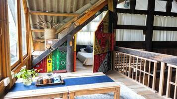 Interior rústico de una cabaña con una escalera de madera, una zona de comedor con mesa y sillas, y un mural de colores en la pared.