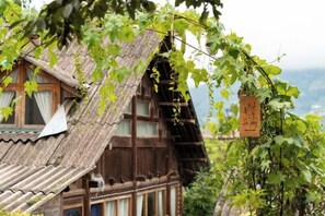 Une maison traditionnelle en bois, dotée d’un toit à pente, entourée de verdure et avec une chaîne de montagnes en arrière-plan.