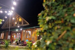 Un edificio rústico de madera con techo a dos aguas, luces colgantes y un pequeño espacio al aire libre con mesas y sillas.