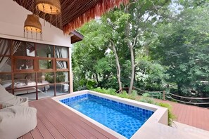 A swimming pool with a wooden deck, a glass-enclosed room with a sofa, and a view of trees. - Villa Cocoon – Jungle River, Villa 29 (Koh Samui)