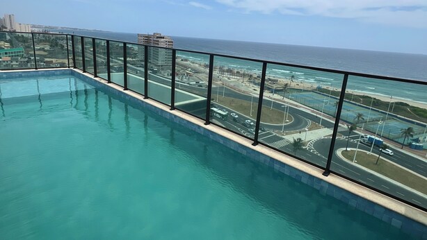 Visão clara de uma piscina com barreira de vidro, com vista para uma área costeira com edifícios e uma estrada.