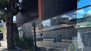 Uma calçada com piso tátil para pessoas com deficiência visual, uma árvore e um edifício com fachada em azulejos.
