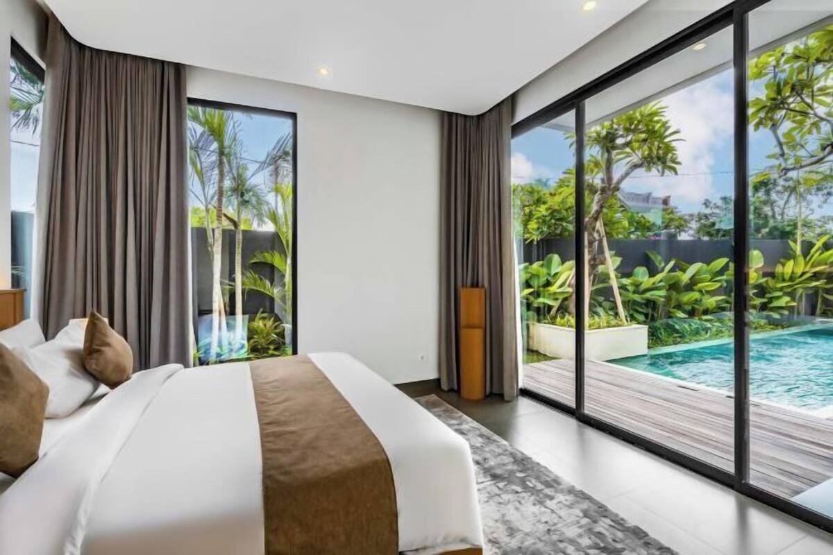 A modern bedroom with a large bed, a view of a swimming pool, and a balcony with greenery.