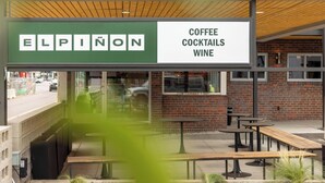 Un espace extérieur aménagé avec des tables et des bancs, une enseigne indiquant « Elpinon : café, cocktails et vins », et un mur de briques en arrière-plan.