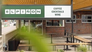 Un espace extérieur aménagé avec des tables et des bancs, une enseigne indiquant « Elpinon : café, cocktails et vins », et un mur de briques en arrière-plan.