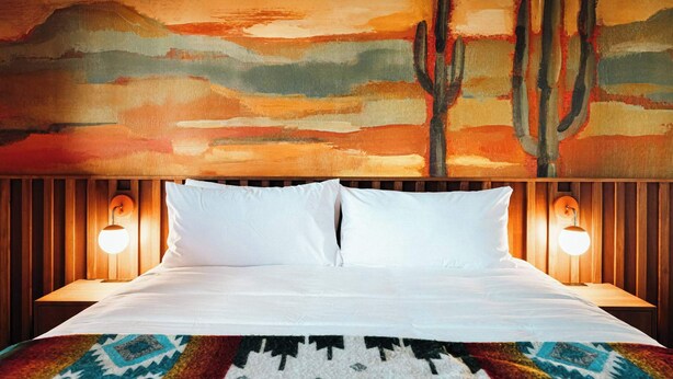 Une chambre d’hôtel avec un lit, deux lampes de chevet, un tapis coloré et un grand tableau représentant un paysage désertique accroché au mur.