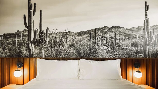 A bed with white linens, two bedside lamps, and a desert-themed mural on the wall behind. - Midnight Cactus Room | La Vista Motel | 203 (Denver)