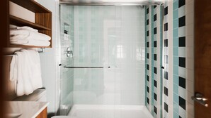 A modern bathroom with a glass-enclosed shower, a wooden shelf with towels, and a tiled wall with a checkered pattern. - Midnight Cactus Room | La Vista Motel | 203 (Denver)