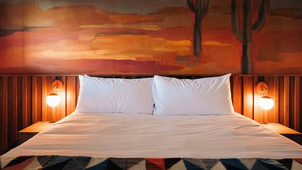 Un lit avec du linge de lit blanc, deux lampes fixées au mur et une fresque représentant un paysage désertique avec un cactus.