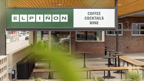 Un espace extérieur aménagé avec des tables et des bancs, une enseigne indiquant « Elpinon : café, cocktails et vins », et un mur de briques en arrière-plan.