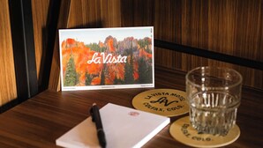 Een houten tafel met een ansichtkaart van La Vista, een glas water, een pen en twee onderzetters met het La Vista logo.