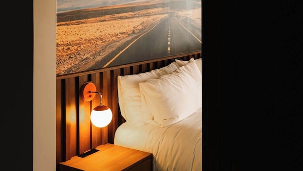 Une chambre avec une grande fresque murale représentant une route de montagne, une tête de lit en bois, une table de chevet avec une lampe et un lit avec une literie blanche.