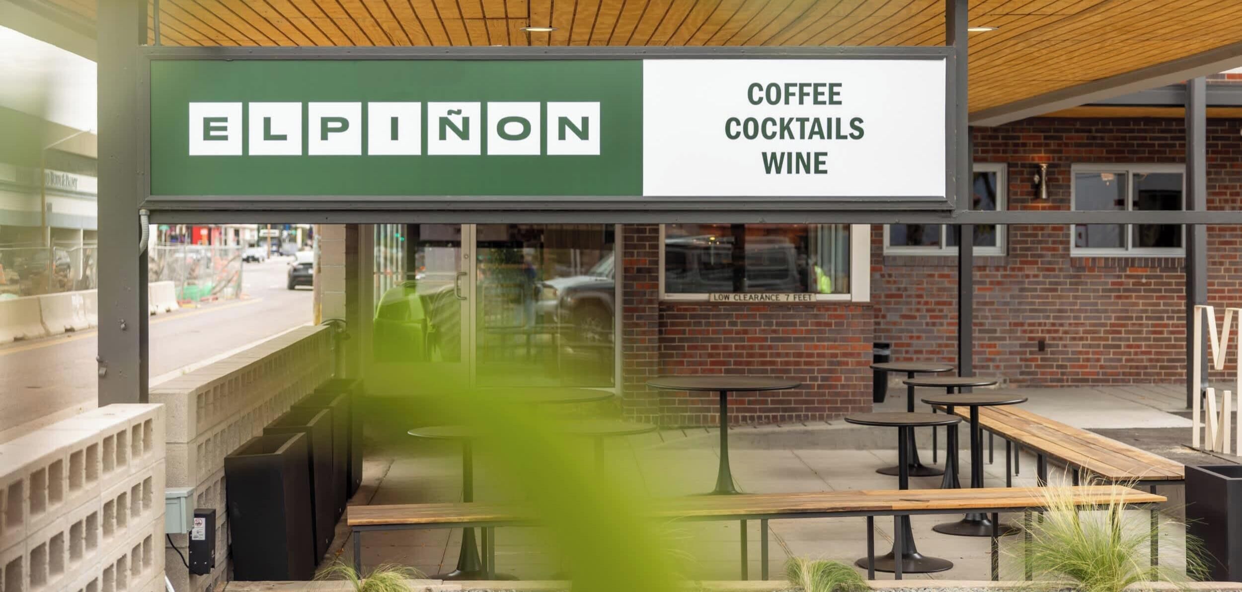 Ett utomhusområde med sittplatser, bord och bänkar, en skylt för Elpinon Coffee, Cocktails and Wine, och en tegelvägg i bakgrunden.
