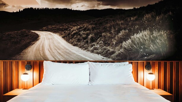 A bed with white linens and two wall-mounted lamps, set against a backdrop of a forested landscape with a winding road. - High Desert Hangout | La Vista Motel | 109 (Denver)