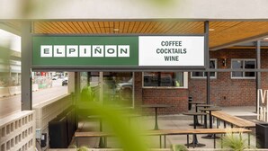 Un área con mesas y bancos para disfrutar al aire libre, un letrero que anuncia Elpinon Coffee, Cocktails y Wine, y un muro de ladrillo al fondo.